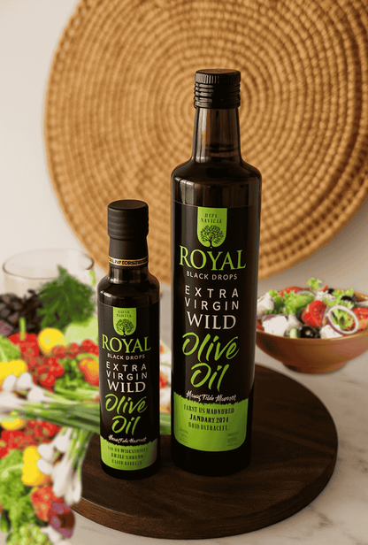 Two bottles of Royal Black Drops Extra Virgin Wild Olive Oil on a wooden board with a woven mat and vegetables in the background.